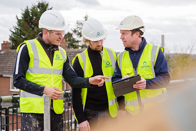 Three LJ Construction workers on a roof top in hard hats and high-visibility jackets reviewing customer relationship management software for a project on a tablet.